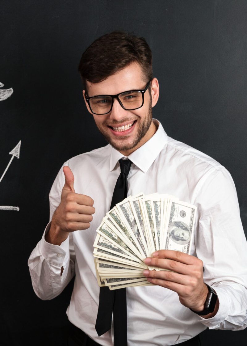 Close-up portrait of young smiling businessman holding bunch of money while showing thumb up gesture, looking at camera, isolated on blackboard with financial statistics